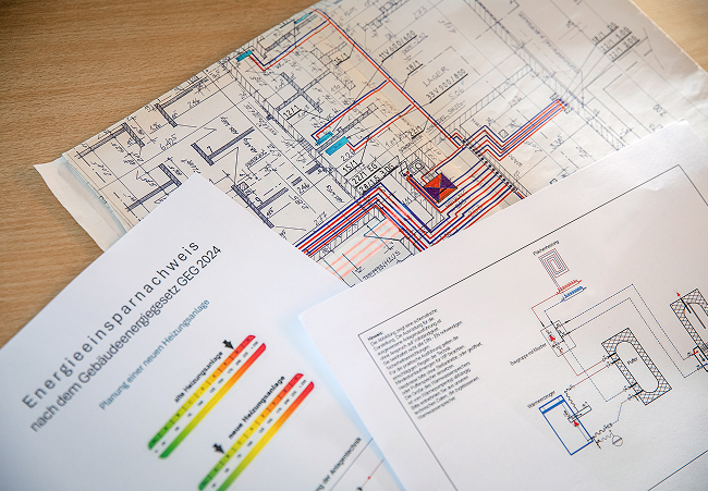 Ein Schreibtisch mit technischen Plänen, Diagrammen und einem Energieeffizienzausweis mit farbigen Energiewerten und deutschem Text, der sich wahrscheinlich auf die Gebäudeplanung und den Energieverbrauch bezieht.