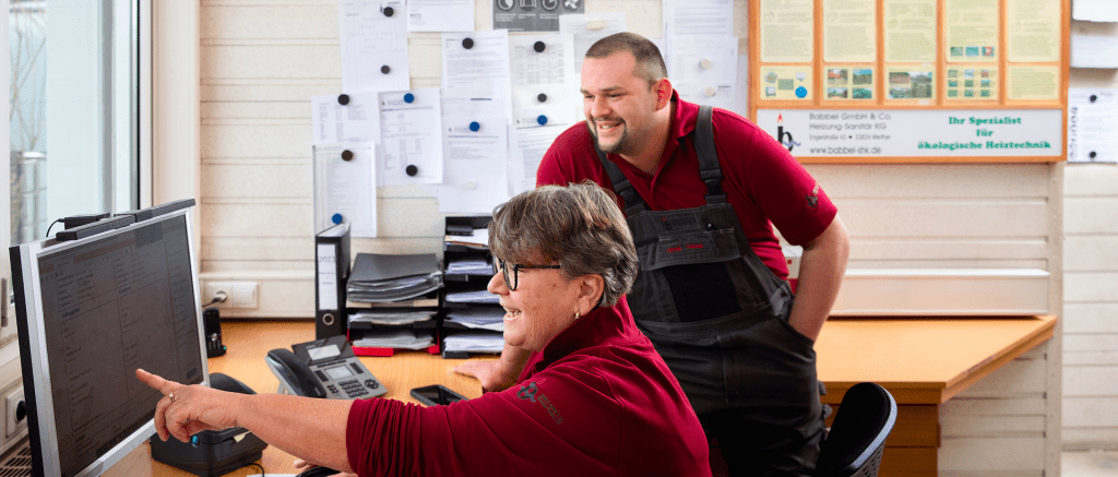 Zwei Personen in kastanienbraunen Hemden arbeiten an einem Schreibtisch in einem Büro. Eine Person sitzt und zeigt auf einen Computerbildschirm, während die andere dahinter steht und lächelt. Papiere und Ordner sind in Regalen geordnet und an die Wand gepinnt. Zwei Personen in kastanienbraunen Hemden arbeiten an einem Schreibtisch in einem Büro. Eine Person sitzt und zeigt auf einen Computerbildschirm, während die andere dahinter steht und lächelt. Papiere und Ordner sind in Regalen geordnet und an die Wand gepinnt.