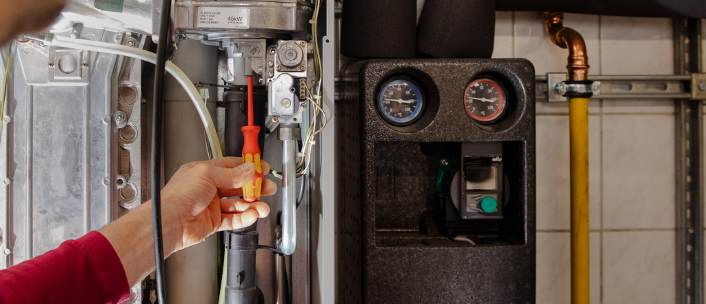 Eine Person benutzt einen rot-gelben Schraubenzieher, um Komponenten in einem Heizkessel oder einer Heizungsanlage einzustellen oder zu reparieren, wobei Rohre, Drähte und Messgeräte in der Nähe sichtbar sind.