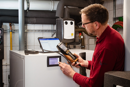 Ein Mann in einem roten Hemd prüft und überwacht mit Hilfe von Diagnosegeräten eine Heizungsanlage in einem Hauswirtschaftsraum, in dessen Nähe Rohre und ein Laptop zu sehen sind.