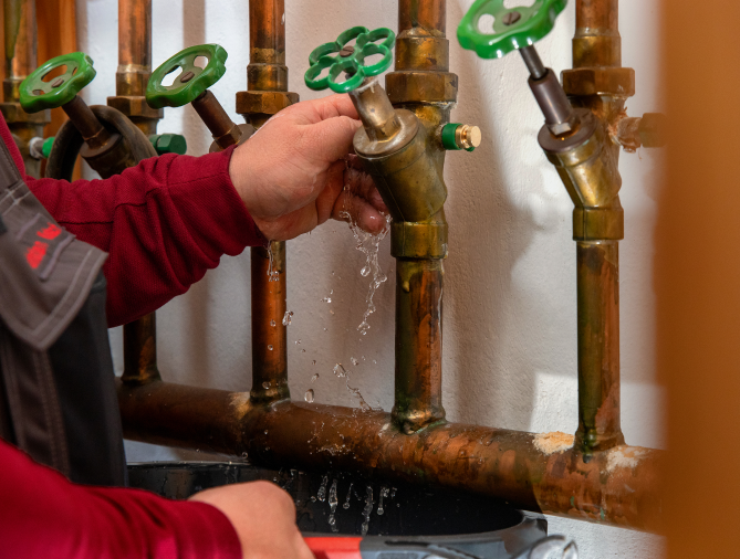 Eine Person in einem roten Hemd öffnet ein Ventil an einem Kupferrohr, woraufhin Wasser herausfließt und spritzt. Andere Rohre und grüne Ventilgriffe sind vor einer weißen Wand zu sehen.