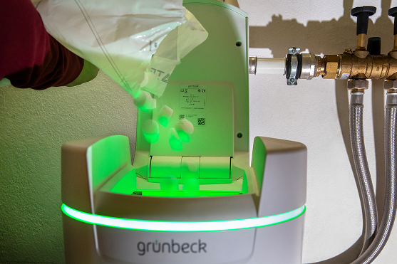 Eine Person schüttet Salztabletten in ein Grünbeck-Wasserenthärtungsgerät, das durch ein grünes Licht beleuchtet wird und an Wasserleitungen an der Wand angeschlossen ist.