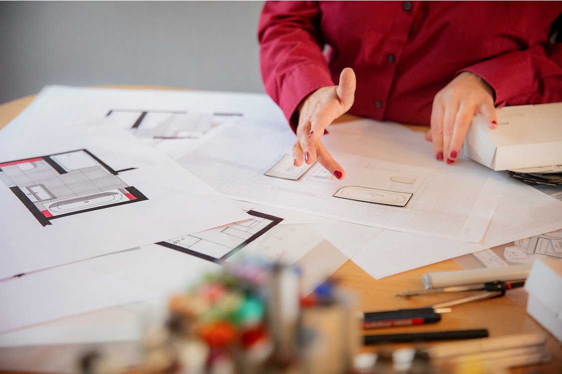 Person in rotem Hemd, die über architektonische Entwürfe und Skizzen gestikuliert, die auf einem Schreibtisch ausgebreitet sind, umgeben von Zeichengeräten und einem Kasten, was auf eine Entwurfs- oder Planungstätigkeit hindeutet.