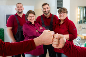 Vier Personen in passenden roten Hemden und schwarzen Overalls stehen lächelnd zusammen im Hintergrund, während zwei Hände im Vordergrund einen Faustschlag ausführen.