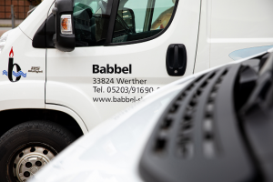 Nahaufnahme eines weißen Lieferwagens mit dem Firmennamen Babbel und Kontaktangaben auf der Tür, teilweise verdeckt durch ein anderes Fahrzeug im Vordergrund.