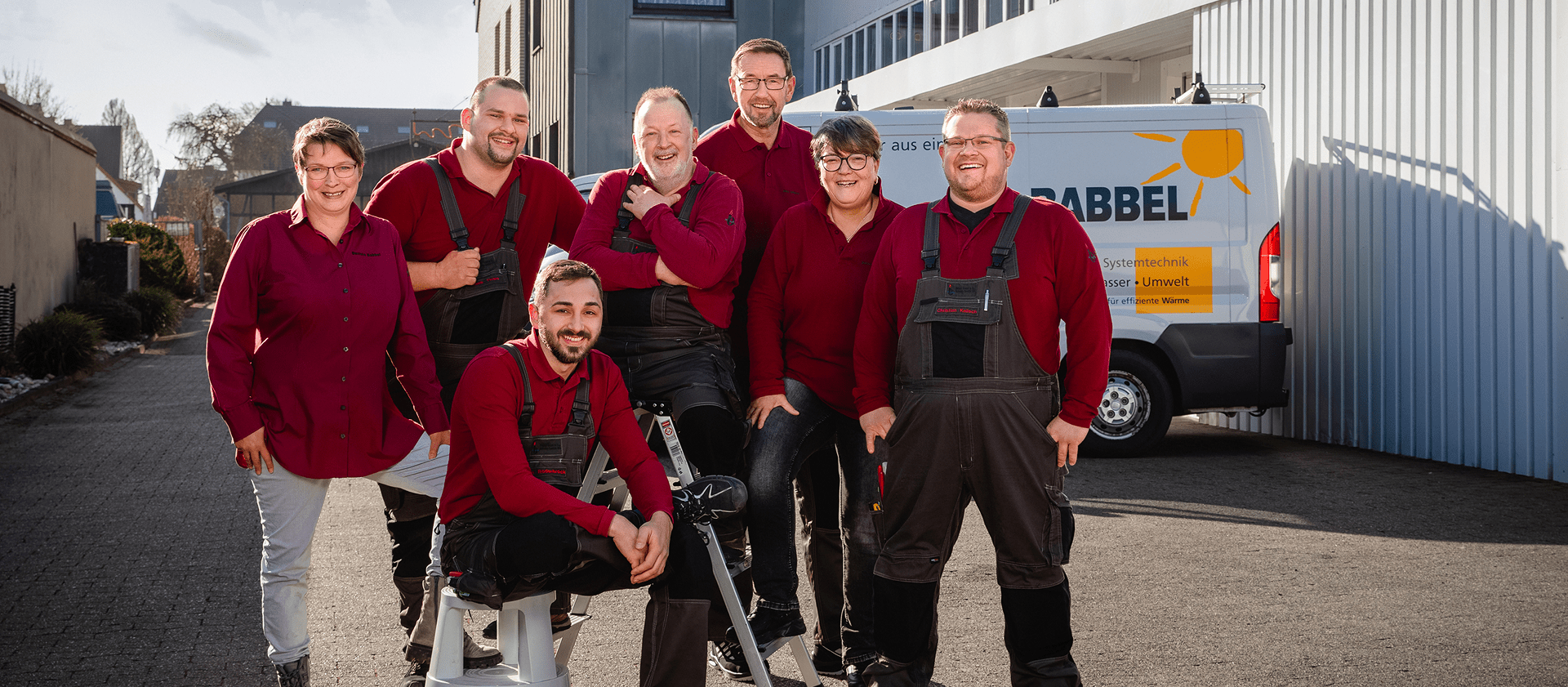 Sieben Personen in passenden kastanienbraunen Hemden und dunklen Overalls lächeln und posieren gemeinsam im Freien vor einem weißen Lieferwagen und einem Gebäude, wobei das Sonnenlicht Schatten auf den Boden wirft. Sieben Personen in passenden kastanienbraunen Hemden und dunklen Overalls lächeln und posieren gemeinsam im Freien vor einem weißen Lieferwagen und einem Gebäude, wobei das Sonnenlicht Schatten auf den Boden wirft.