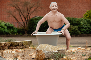 Eine lebensgroße Statue eines lächelnden älteren Mannes mit Schnurrbart und blauer Badehose, der in einer Badewanne im Freien steht, umgeben von Felsen und Grünpflanzen.